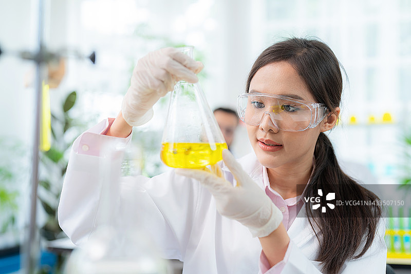 年轻的亚洲研究员穿着实验服，在烧瓶中摇匀溶液。聪明的女人在现代实验室从事医学和生物技术工作。生物植物概念的研究与发展图片素材