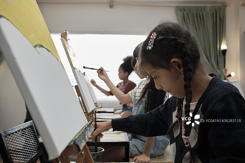 在美术教室里，一个小女孩正专注地在画布上画亚克力彩画。图片素材