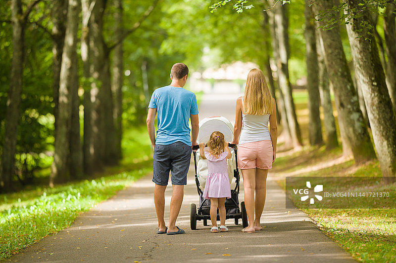 在温暖阳光明媚的夏日，年轻的父母和小女儿推着婴儿车走在公园的树巷里。共度时光，呼吸新鲜空气。两个孩子的家庭。后视图。图片素材