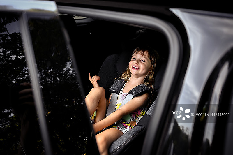 一个坦率的无忧无虑的蹒跚学步的女儿坐在车里准备旅行的肖像。发短信的地方。图片素材