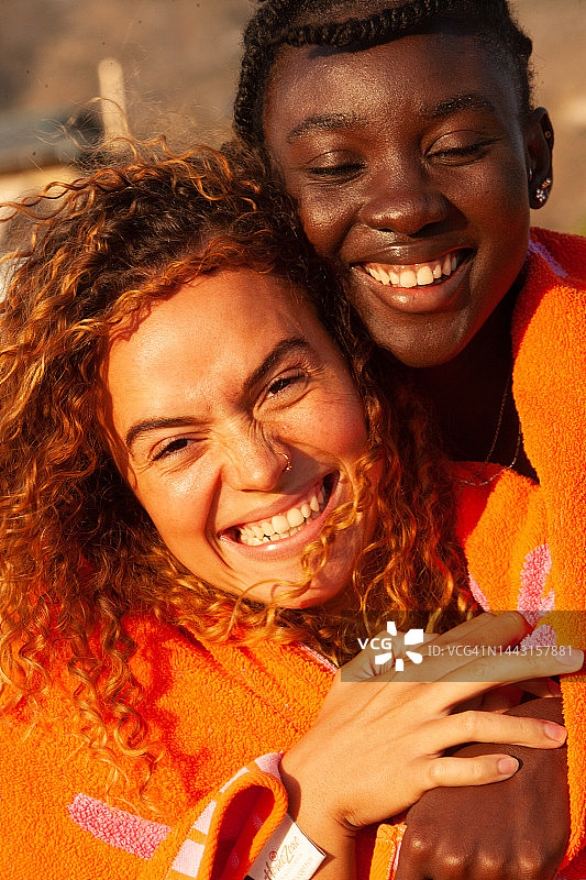 两个狂喜的美丽的年轻女子看着镜头，两个二十多岁的女孩和那里的女朋友与自然辫子的afro和覆盖着橙粉色的毛巾覆盖他们是她的朋友长卷金色头发的后面图片素材