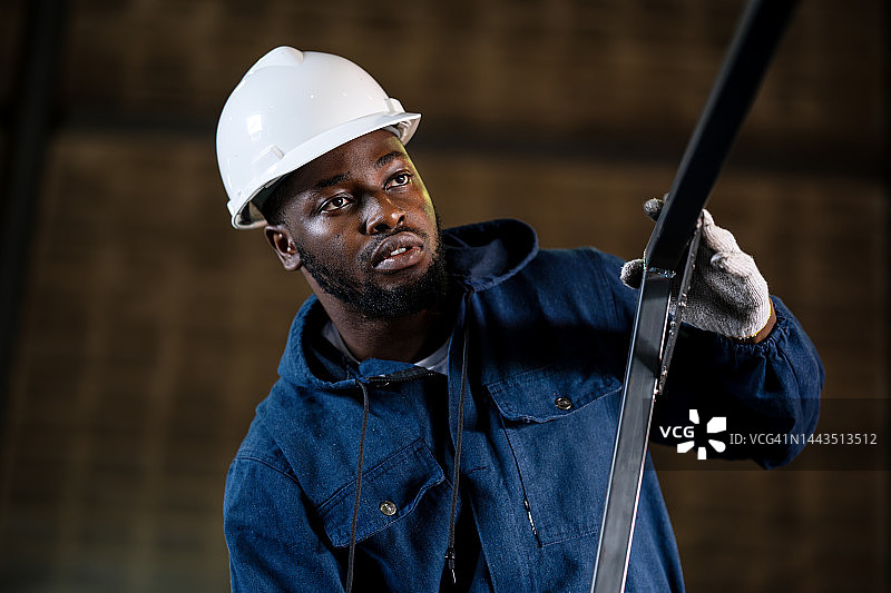 横角焊接强度。一名非裔美国焊工在工厂焊接期间检查管架零件表面精度的低角度视图。图片素材
