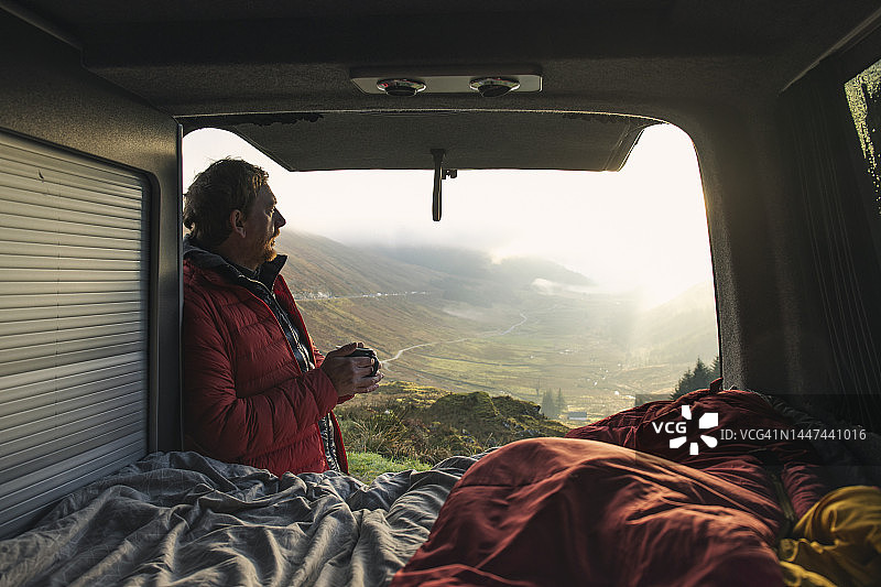 一个成熟的男人坐在他的露营车后面，一边喝着热饮，一边欣赏美丽的晨光图片素材