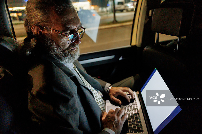 成熟的商人在晚上开车时坐在汽车后座上使用笔记本电脑图片素材