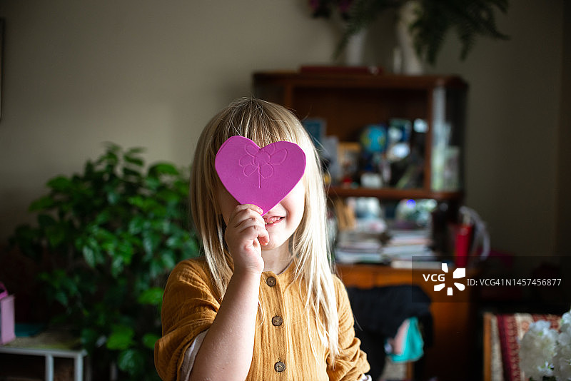 一个穿着橙色连衣裙的可爱快乐的小女孩，手里拿着用纸剪成的粉红色心形，对着镜头笑着、高兴着。图片素材