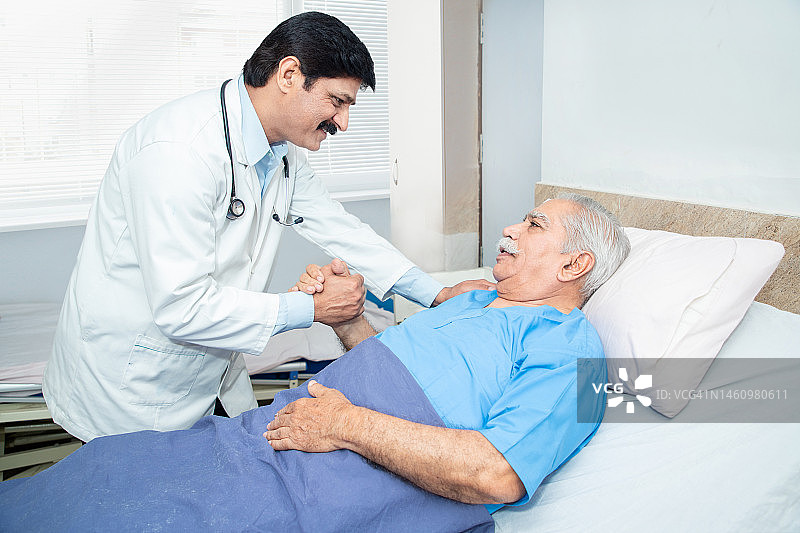 在诊所或医院，微笑的印度医生把手放在躺在床上的老年男性病人的肩膀上。老年人保健理念。图片素材
