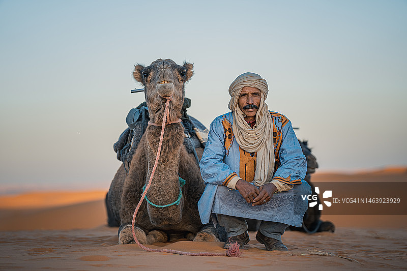 摩洛哥骆驼司机蹲在撒哈拉沙漠与骆驼看着相机图片素材