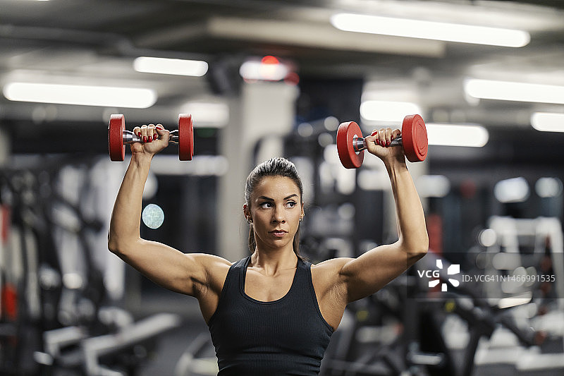 一个强壮的肌肉女健美运动员在健身房举哑铃的肖像。图片素材