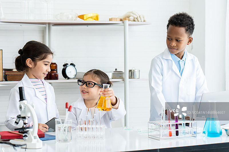 在化学教室里，一小群学生穿着白色的实验大褂，在架子和桌子上放着许多实验工具。一个戴眼镜的小女孩在帮助实验时，手里拿着黄色的化学瓶。图片素材