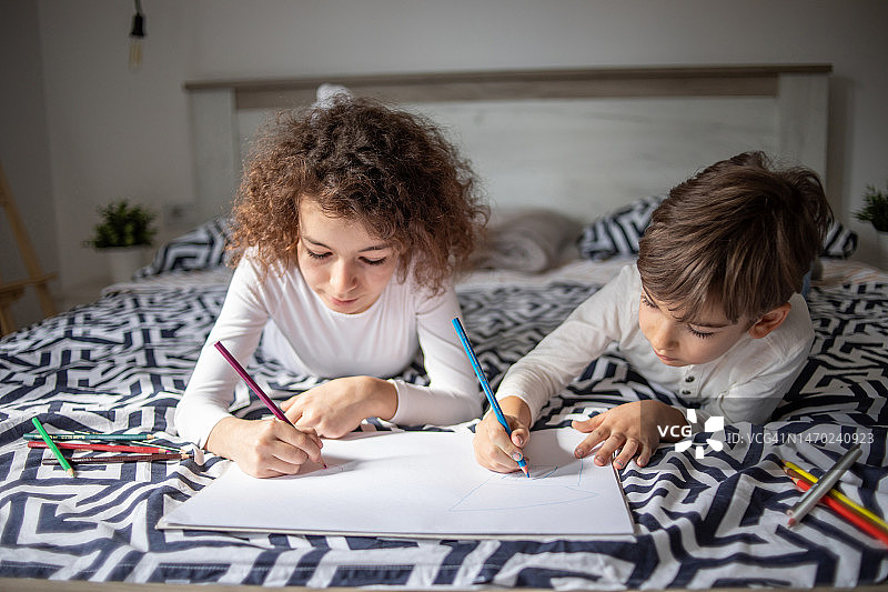两个孩子，弟弟和妹妹，在卧室的床上用木笔在纸上画画图片素材