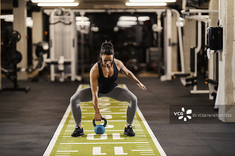 一位强壮的女健身运动员正在健身房里蹲着举壶铃。图片素材