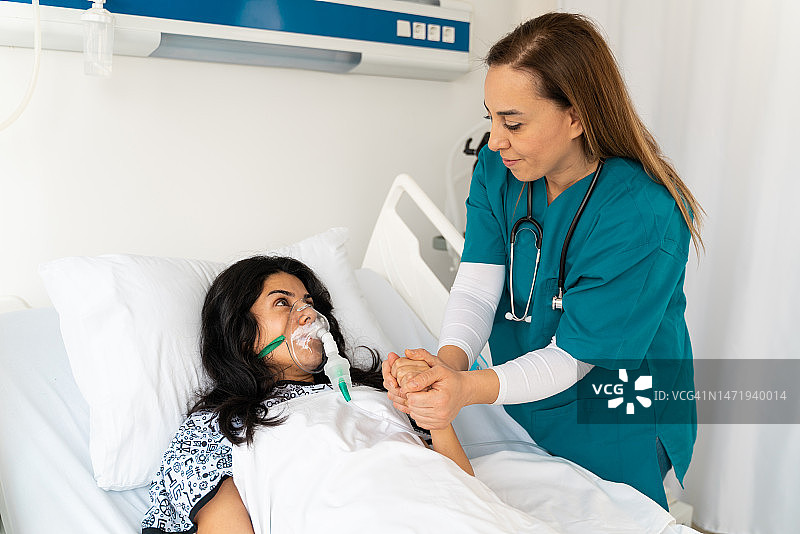 一名妇女戴着氧气面罩躺在病床上，与关怀的医生或护士手牵手图片素材
