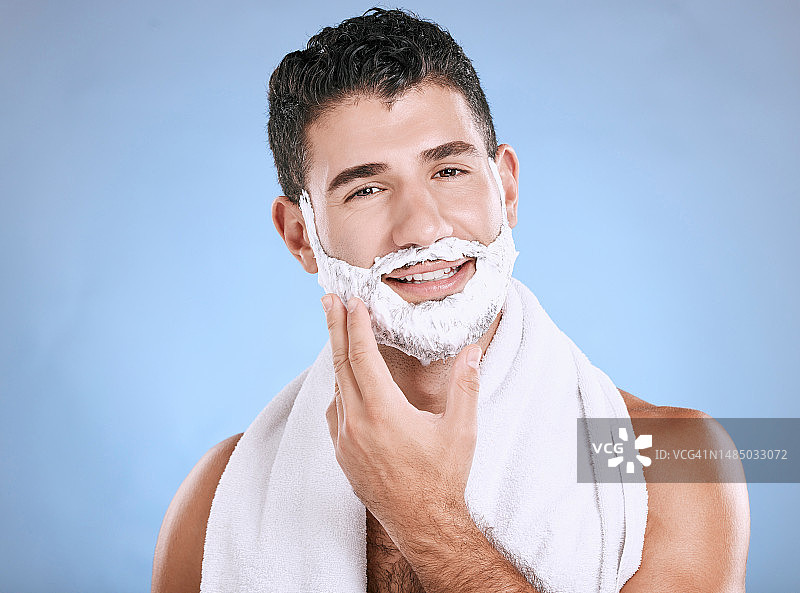 剃须泡沫在胡须上，微笑和手在脸上用毛巾和产品植入在工作室模拟。剃须膏，面部和头发或皮肤护理的快乐男模美容，孤立的蓝色背景。图片素材