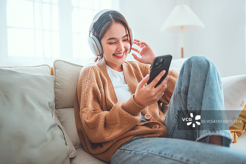 快乐的亚洲女人坐在家里的沙发上听着手机里的音乐，微笑的女孩在早上戴着耳机放松，是时候放松了。图片素材