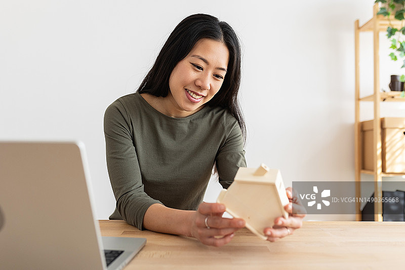 微笑着的年轻亚洲女人拿着笔记本电脑坐在家里的办公室里，手里拿着一个小木屋模型，希望将来有自己的家。图片素材