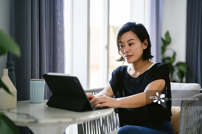年轻的亚洲女性坐在靠窗的桌子上，在家工作时使用数码平板电脑。家庭办公室图片素材