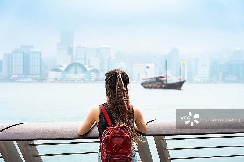 一位年轻的亚洲女游客微笑着眺望维多利亚港海滨长廊，享受着宁静的空气和壮丽的城市景观。图片素材
