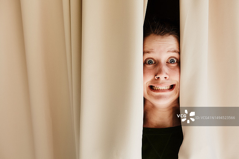 一位年轻女子透过窗帘向外看，脸上露出恐惧的表情图片素材