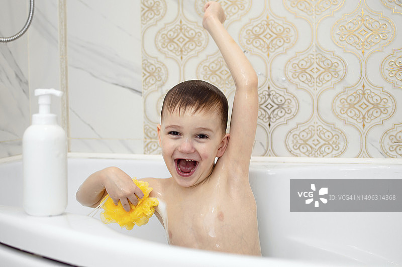 有趣的孩子正在洗澡，坐在有肥皂和洗发水泡沫的白色浴缸里，用他的头发洗他的头。儿童用洗发水、护发产品和肥皂。健康和卫生的概念图片素材