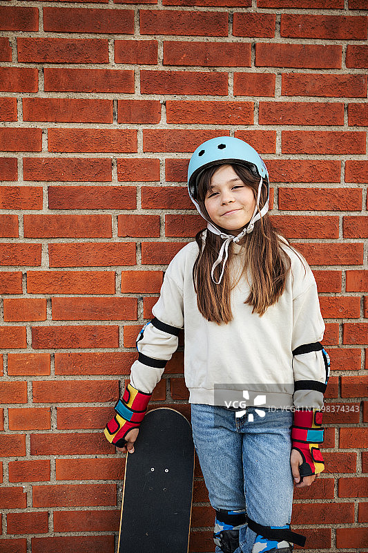 一个快乐的小女孩抱着滑板，靠着砖墙。图片素材