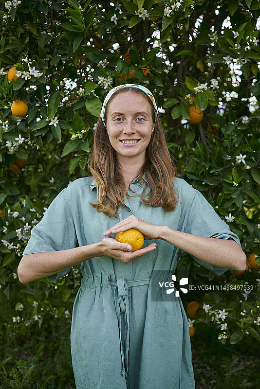 在果园的树前，微笑的女人拿着新鲜的橘子图片素材