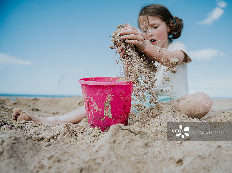 一个小女孩从海滩上舀了一把沙子，放进她的粉色塑料桶里图片素材