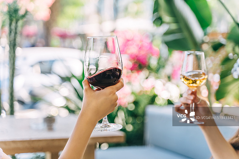在一个阳光明媚的夏日，两只手拿着红葡萄酒和白葡萄酒碰杯。图片素材