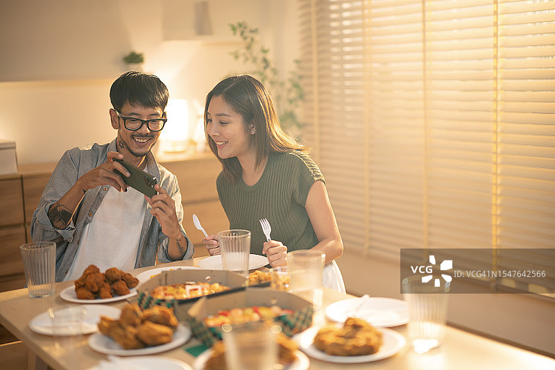 用科技加强家庭关系。一名东南亚男性在晚宴上向女友展示照片，餐桌上摆放着巨大的披萨和炸鸡或意大利菜。图片素材