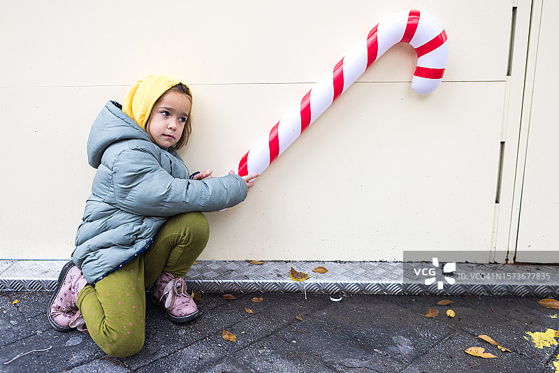 小女孩戴着兜帽，穿着大衣，拿着一根巨大的白色糖果手杖，上面有红色的圣诞条纹，作为装饰，挂在街上的彩色墙上。圣诞概念，装饰，装饰，传统。图片素材