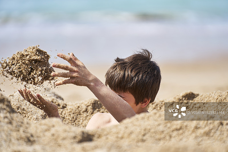 一个男孩被埋在比亚里茨海滩上的沙子里，他穿着泳衣，正对着大海图片素材