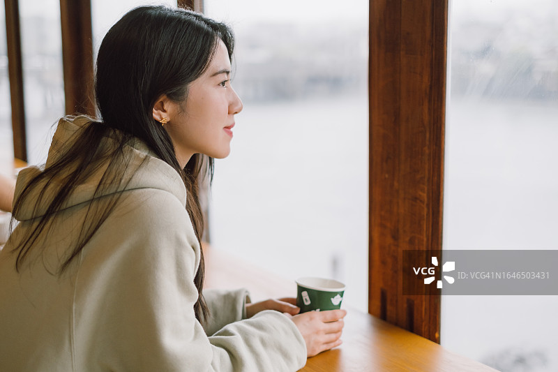 在宇治的日本咖啡馆喝酒的年轻亚洲女子侧视图，欣赏窗外的风景。享受下雨天放松的记忆。食物观念，外出就餐，旅游和度假。图片素材