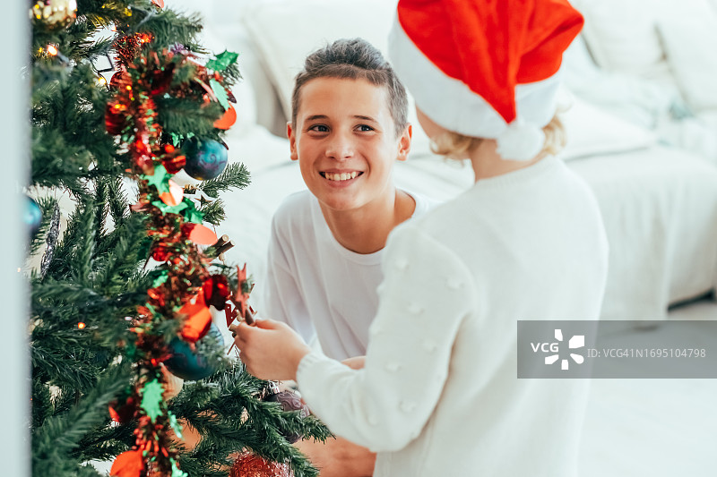 十几岁的小弟弟和可爱的小妹妹戴着红色的圣诞帽在家里装饰圣诞树。寒假快乐视频。图片素材