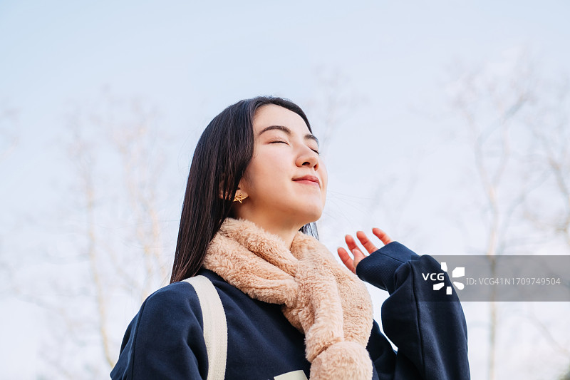 年轻的亚洲女子在冬天晴朗的蓝天下闭着眼睛呼吸着清凉的空气。在大自然中享受清新的空气。可持续的生活方式。无忧无虑，自由自在。与自然的概念联系起来。图片素材