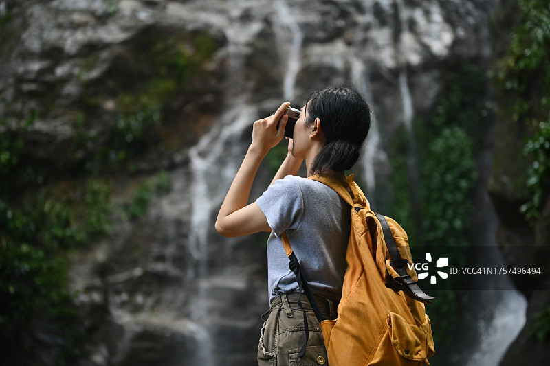 年轻女子背包客站在瀑布前，用复古相机拍照图片素材