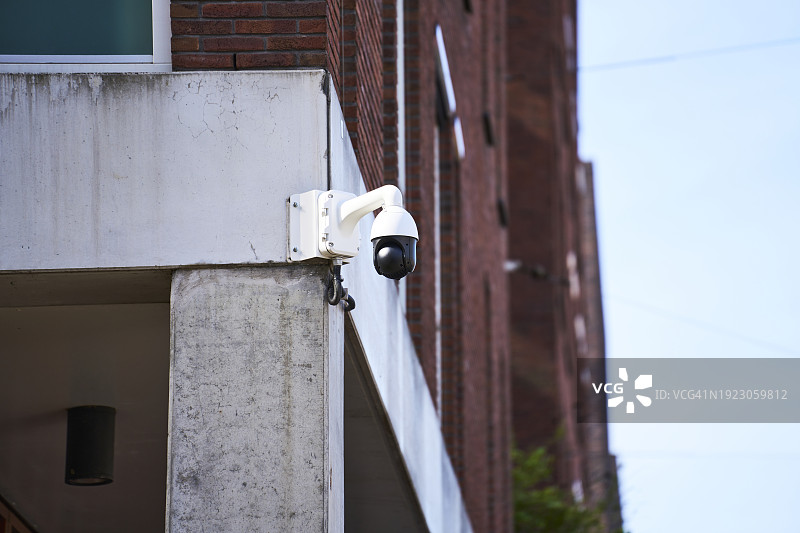 一个安全摄像头，正面视图，放置在一个大建筑物的角落里，背景是红砖墙，另一个建筑物的侧面有些模糊。图片素材