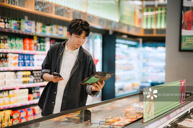 亚洲中国学生在超市选购商品时看智能手机。图片素材