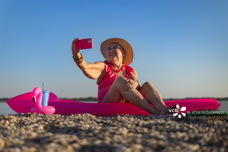 一位老妇人手持智能手机，坐在粉红色的泳池浮子上，在沙滩上自拍。一位老年妇女在海边享受暑假。退休后旅行的概念。图片素材