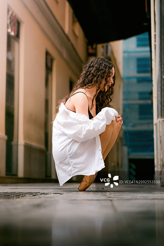 在城市街道上，一名芭蕾舞女演员以一种具有挑战性的姿势蹲下脚尖休息。图片素材