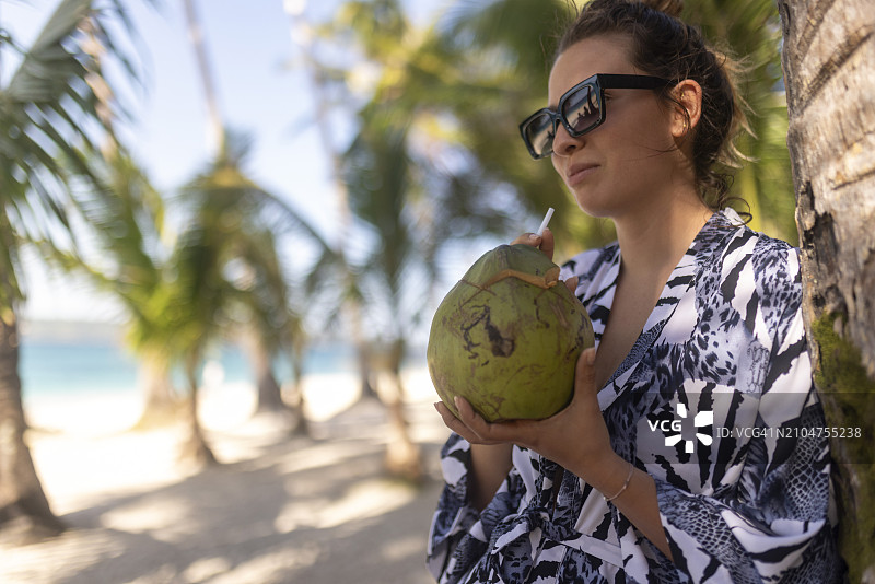 一个微笑的女人站在沙滩上，喝着鸡尾酒，享受着周围的美景图片素材