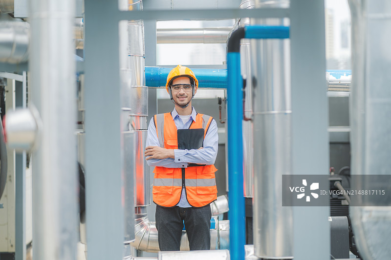 穿着安全装备的工人自信地站在工业设施内，背景是机械。图片素材