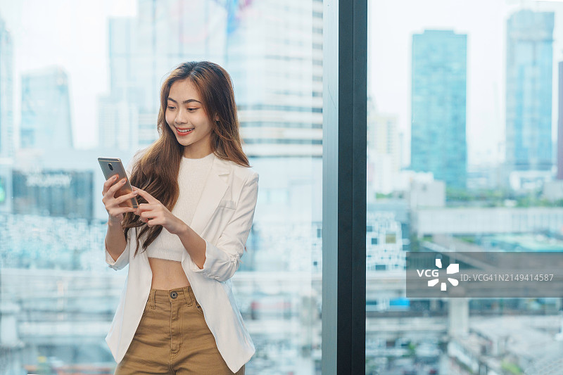 年轻的专业亚洲商务女性穿着西装，手持无线耳机，手持智能手机，用手机在城市进行视频通话。图片素材