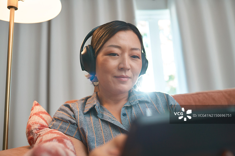 音乐耳机，平板电脑和亚洲女人在家里客厅的沙发上进行在线学习或语言学习。收音机，流媒体和技术与人听音频或声音在公寓图片素材