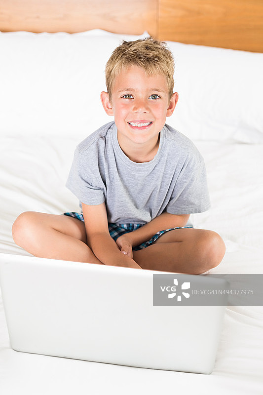 可爱的金发男孩坐在床上用笔记本电脑图片素材