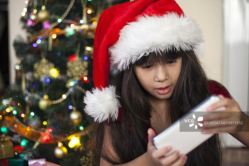 亚洲女孩戴着圣诞老人的帽子，拿着圣诞礼物——智能手机图片素材