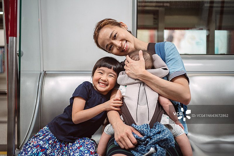 漂亮的年轻妈妈和她可爱的小女儿和婴儿在婴儿车里坐在火车上，对着镜头开心地微笑。图片素材
