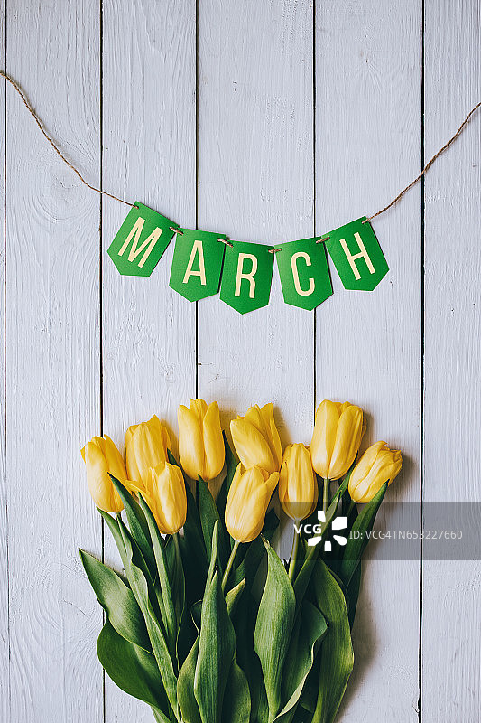 黄色的郁金香束在白色的木板上乡村谷仓乡村桌子的背景。三月绿花环纸刻字、文字、字母、题词。美丽的垂直的明信片。图片素材