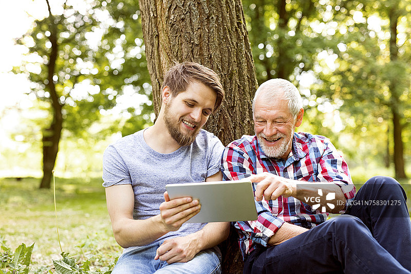 快乐的老父亲坐在他的成年儿子在一个公园看平板电脑图片素材