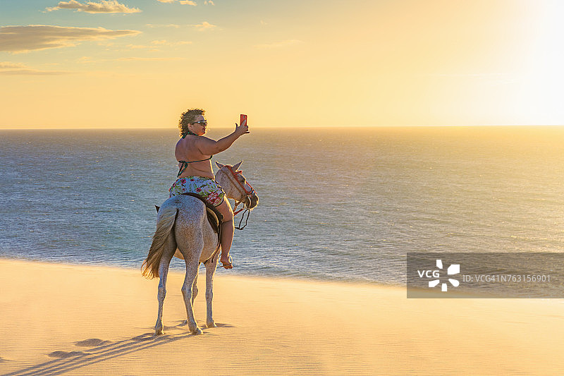 南美巴西塞阿拉岛的Jericoacoara，一名女子在沙滩上骑马，用智能手机拍摄风景图片素材