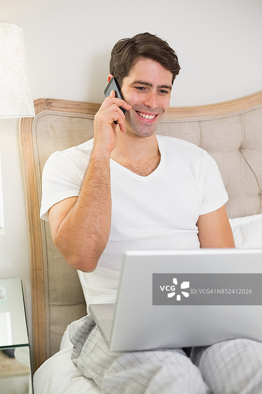 随意微笑的男人在床上使用手机和笔记本电脑图片素材