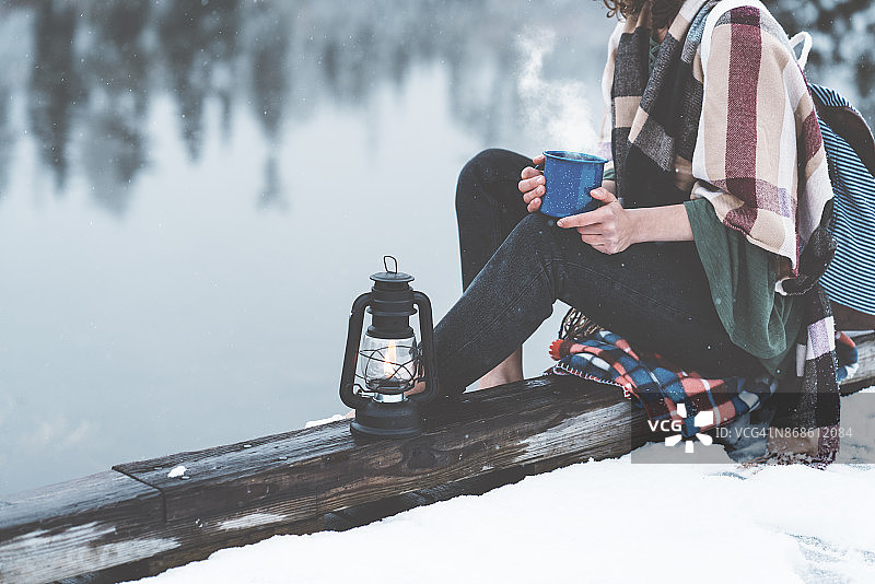 拿着热饮料和油灯的女人坐在白雪覆盖的湖边图片素材